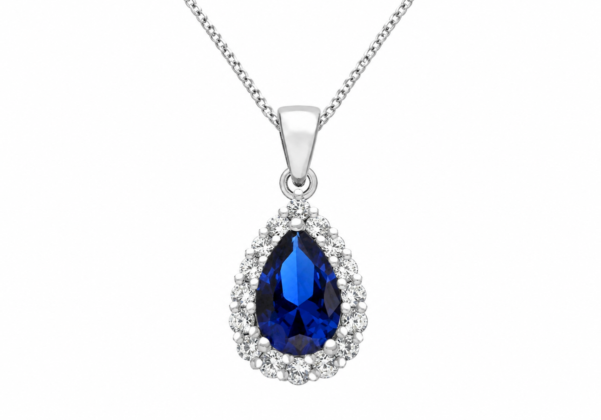 A silver pendant necklace featuring a teardrop-shaped deep blue gemstone surrounded by a halo of small clear stones, suspended from a silver chain on a white background.